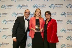 Three people smile at the camera. The man on the left is bald, has glasses and is wearing a black tux, the lady in the middle is strawberry blonde and wearing an orange blazer and holding a trophy, and the lady on the right is wearing a red blazer and has short brown hair and glasses. They are standing in front of a Chemistry Australia banner.