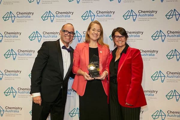 Three people smile at the camera. The man on the left is bald, has glasses and is wearing a black tux, the lady in the middle is strawberry blonde and wearing an orange blazer and holding a trophy, and the lady on the right is wearing a red blazer and has short brown hair and glasses. They are standing in front of a Chemistry Australia banner.