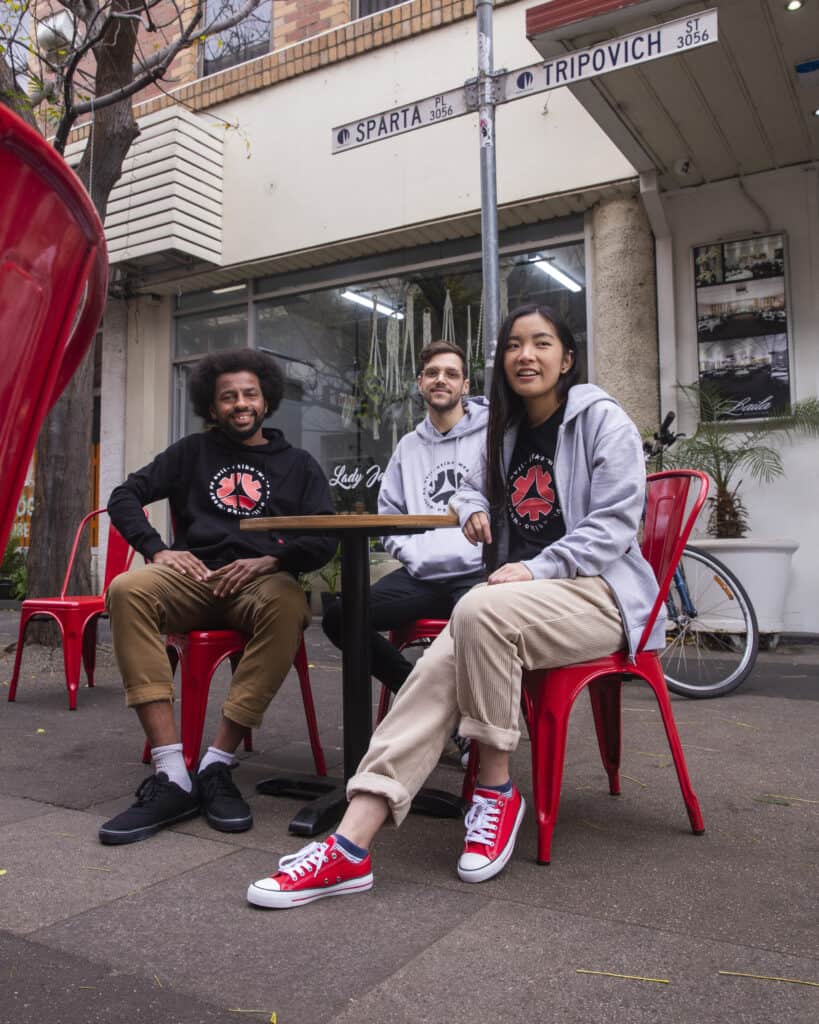 A group of young people (two men and a woman) sitting at a red cafe table on a street, wearing Etiko branded streetwear.