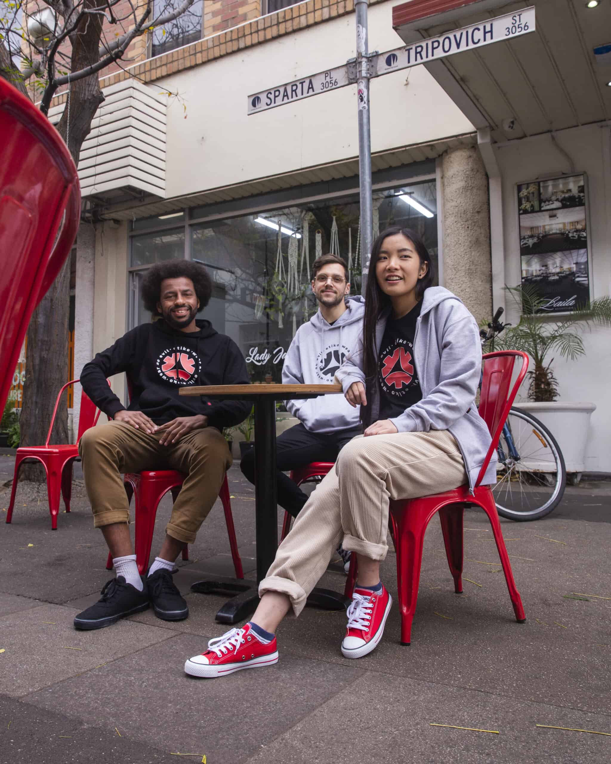 A group of young people (two men and a woman) sitting at a red cafe table on a street, wearing Etiko branded streetwear.