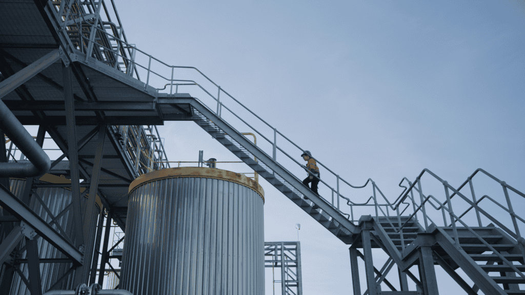 A woman in a fluoro tradie jumper walks up a talk ladder between silos. She is silhouetted against the sky.