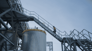 A woman in a fluoro tradie jumper walks up a talk ladder between silos. She is silhouetted against the sky.