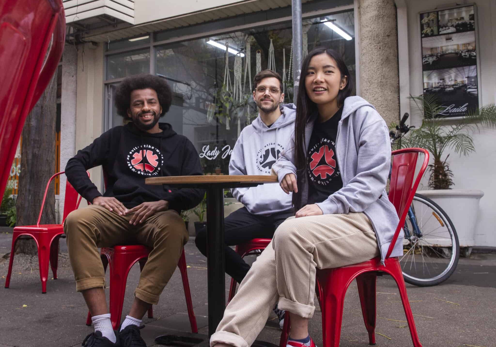 A group of young people (two men and a woman) sitting at a red cafe table on a street, wearing Etiko branded streetwear.