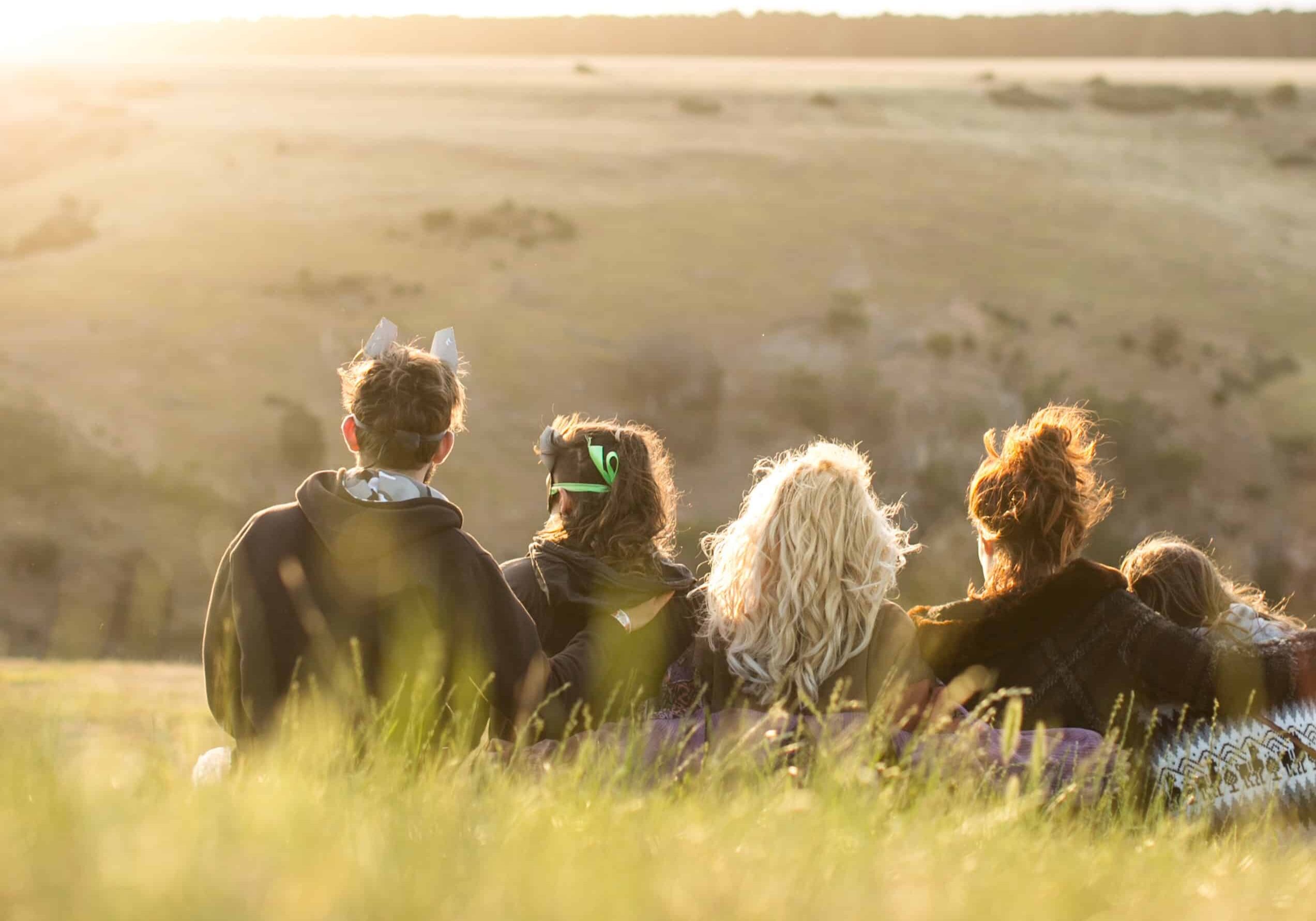 Five young people sit on a grassy hill watching the sun set over fields.