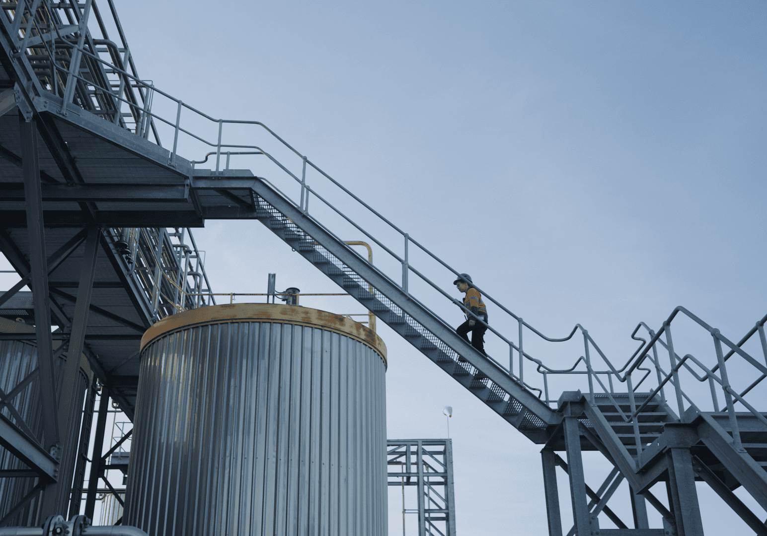 A woman in a fluoro tradie jumper walks up a talk ladder between silos. She is silhouetted against the sky.