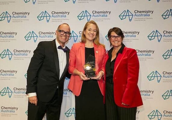 Three people smile at the camera. The man on the left is bald, has glasses and is wearing a black tux, the lady in the middle is strawberry blonde and wearing an orange blazer and holding a trophy, and the lady on the right is wearing a red blazer and has short brown hair and glasses. They are standing in front of a Chemistry Australia banner.